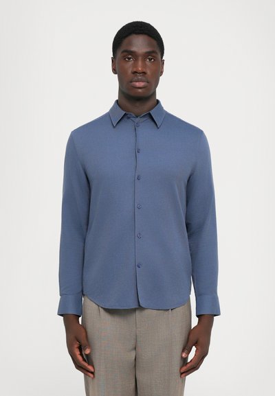 Blauwe langarmsgemouwen shirt met een knoopsluiting aan de voorkant, puntige kraag en afgeronde zoom; gemaakt van een gestructureerde stof. Draagt men met lichtgrijze broeken.