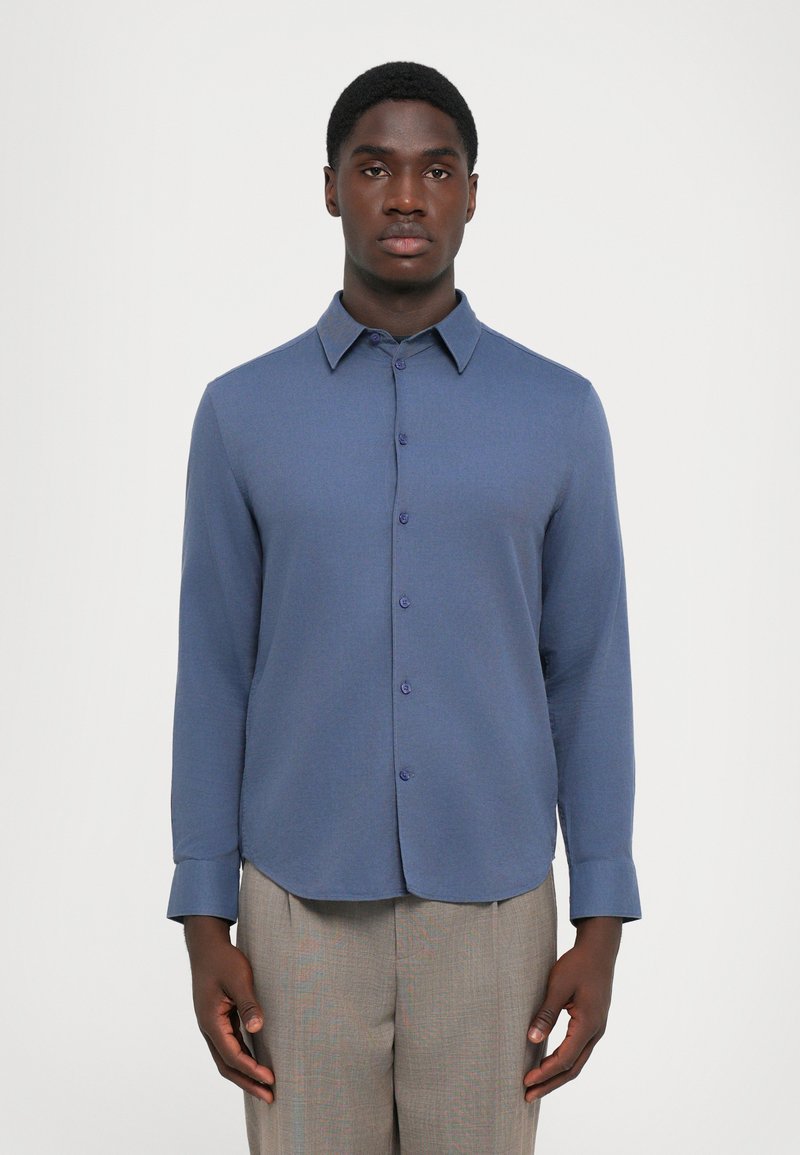 Blauwe langarmsgemouwen shirt met een knoopsluiting aan de voorkant, puntige kraag en afgeronde zoom; gemaakt van een gestructureerde stof. Draagt men met lichtgrijze broeken.