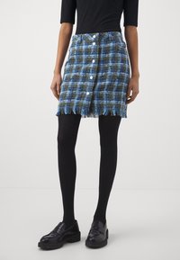 Jupe courte à carreaux aux tons bleus et verts, avec une patte de boutonnage sur le devant et un ourlet effiloché. Portée avec des collants noirs et des mocassins, offrant un look superposé.