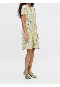 Robe portefeuille fleurie à manches courtes, avec des fleurs jaunes et bleues sur fond crème, jupe à volants. Associée à des talons verts à brides.