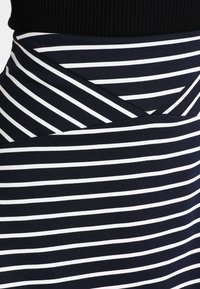 Jupe rayée bleu marine et blanche avec un design élégant et ajusté. Elle possède un détail de taille croisée et une ceinture en ribbed noir. Texture de tissu douce.