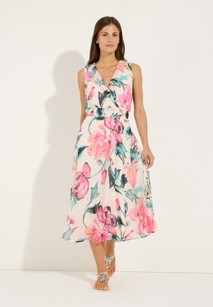 Frau in ärmellosem weißen Kleid mit rosa und grünem Blumenmuster, barfuß laufend mit silbernen Sandalen mit Schmucksteinen vor einfachem weißem Hintergrund.