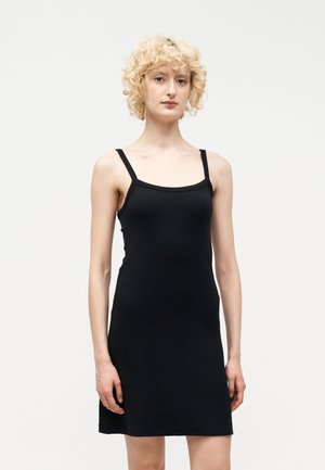 Νεαρό άτομο με κοντά σγουρά ξανθά μαλλιά που φοράει αμάνικο μαύρο φόρεμα με λεπτές τιράντες, στέκεται μπροστά σε ένα απλό φωτεινό φόντο.