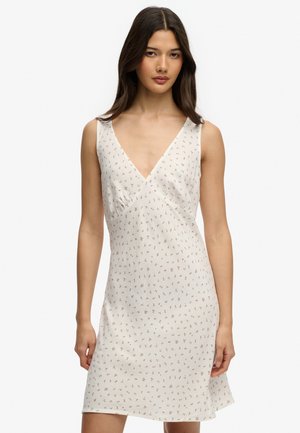 Frau trägt ein ärmelloses, weißes V-Ausschnitt-Kleid mit einem kleinen beigen Blumenmuster, stehend vor einem schlichten weißen Hintergrund.