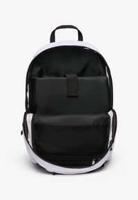 Sac à dos avec un intérieur en tissu noir, un contour extérieur blanc, une fermeture éclair, et plusieurs compartiments y compris une poche en mesh et une sangle ajustable.