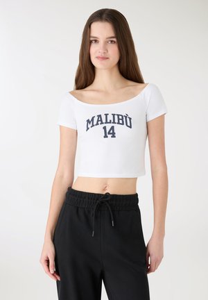 T-shirt bianca corta con la scritta "MALIBÙ 14" in blu navy, caratterizzata da un design a spalle scoperte. Abbinata a pantaloni della tuta neri con coulisse.