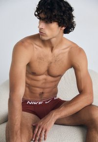 Red boxer briefs with a white Nike logo on the waistband. The model has curly hair and a muscular build, seated against a textured background.