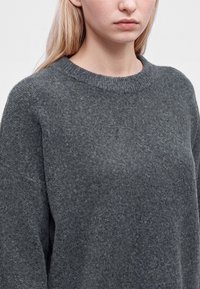 Personne blonde portant un pull en maille texturée gris foncé avec un col rond, visible du cou jusqu'au milieu du torse sur un fond clair.