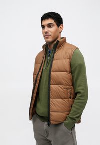 Barna pufi mellény cipzáros elülső résszel és magas gallérral, zöld pulóver fölött rétegezve. Steppelt mintázattal és oldalzsebekkel rendelkezik.