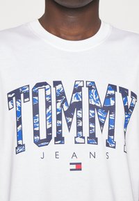 Bílý bavlněný tričko s textem "TOMMY" v modrém a bílém horském vzoru, s nápisem "JEANS" níže a akcentem loga vlajky Tommy.