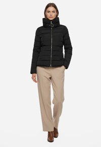 Giacca a piuma nera con cuciture verticali, colletto alto e fibbia regolabile. Presenta una zip frontale e tasche laterali. Abbinata a pantaloni beige.