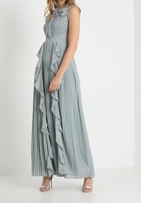 Vaaleansininen, hihaton maksimekko, jossa on poimutuksia ja röyhelöitä sivussa. Kiinteä bodice ja valuva hame. Käytössä hopeiset korokepohjasandaalit.