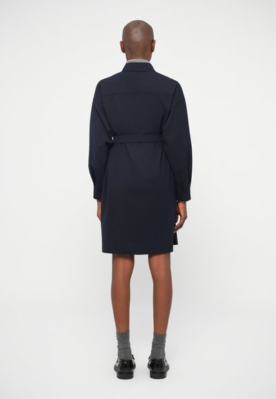 Μπλε ναυτικού φόρεμα με ζώνη στη μέση, γιακά και μακριά μανίκια. Φτιαγμένο από λείο ύφασμα, μεσαίου μήκους, με πλάγιες σχισμές και χαλαρή εφαρμογή.