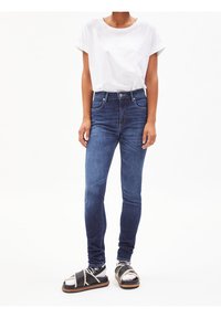 Modré džíny s vysokým pasem, úzkého střihu, s hladkým povrchem a jemnými detaily šití, v kombinaci s jednoduchým bílým tričkem a černými sandály.