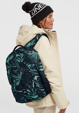 Navy backpack with green palm leaf pattern, padded straps, front pocket, and mesh side pocket; worn by a person in a cream-colored jacket.