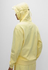 Persona che indossa una felpa gialla chiaro con cappuccio regolabile, vista da dietro su uno sfondo grigio uniforme.