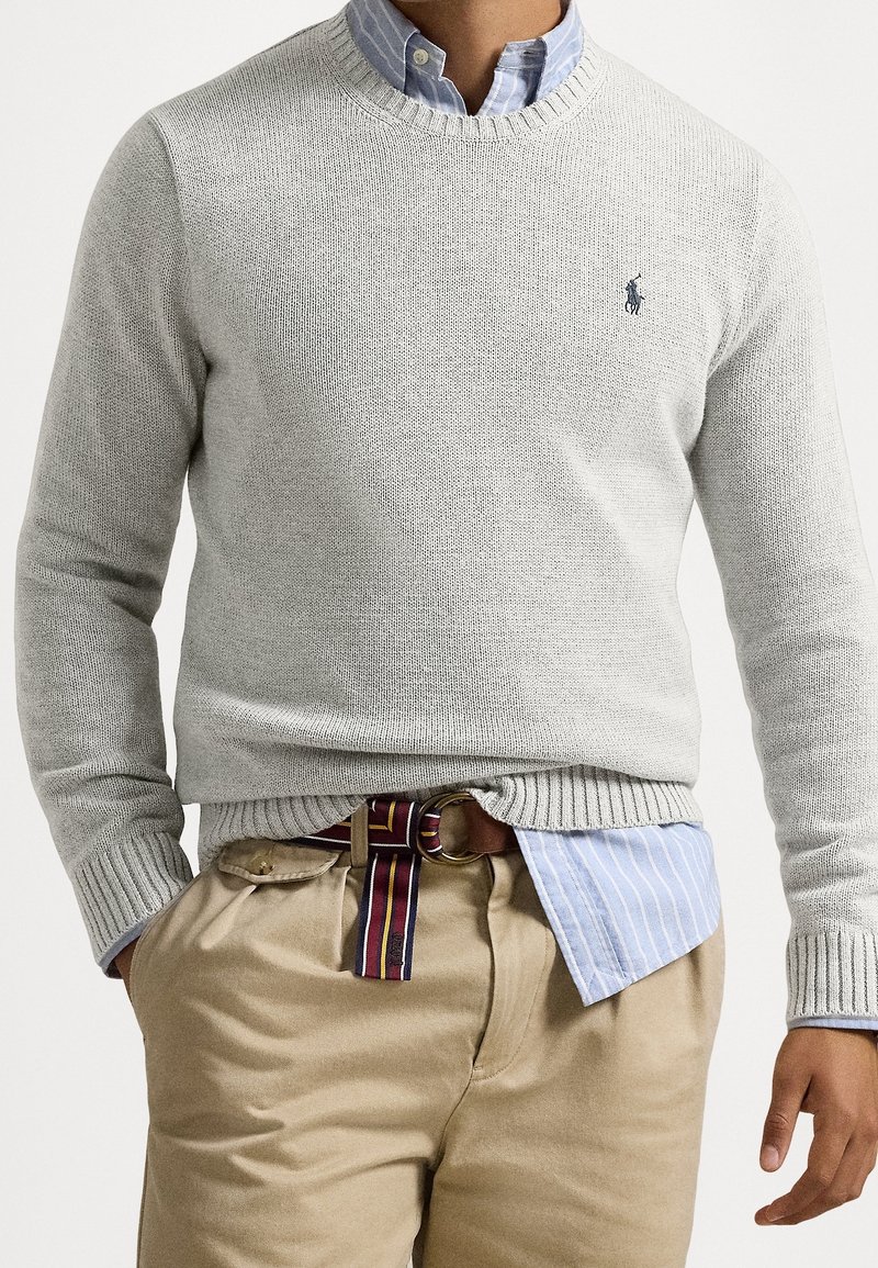 Homme portant un pull en maille gris clair par-dessus une chemise à rayures bleues à boutons, pantalon beige et ceinture en tissu rayé, les mains dans les poches.