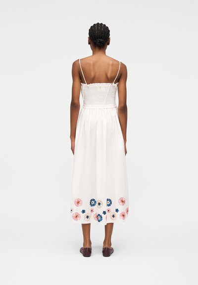 Femme aux cheveux tressés portant une robe midi blanche sans manches avec une broderie florale à l'ourlet, debout pieds nus sur un fond uni.