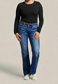 Personne portant un haut noir à manches longues, un jean bleu droit et des baskets blanches, debout devant un fond beige uni.