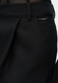 Κοντινό πλάνο μαύρου ραμμένου παντελονιού με ψηλό μέση, θηλιά ζώνης, μπροστινή πιέτα και μεταλλική ετικέτα με την επωνυμία κοντά στην τσέπη.