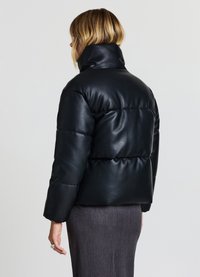 Giacca a vento nera con colletto alto, realizzata in materiale sintetico. Presenta un design trapuntato e un taglio corto, indossata sopra una gonna grigia.