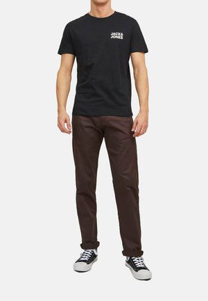Schwarzes Baumwoll-T-Shirt mit weißem "JACK & JONES"-Logo auf der linken Brust, kombiniert mit braunen Hosen und schwarzen Sneakern mit weißen Sohlen.