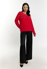 Maglione rosso lavorato a maglia con dettagli in perle vicino allo scollo, abbinato a pantaloni neri a vita alta e a gamba larga, e tacchi neri.