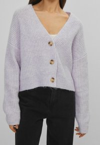 Lilla strikket cardigan med V-udskæring, fire store knapper, cropped design og brede ærmer, parret med sorte højtaljede bukser.