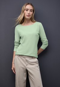 Lichtgroene blouse met lange mouwen in zachte stof en een bootnek, gecombineerd met beige op maat gemaakte broeken met een subtiele textuur.