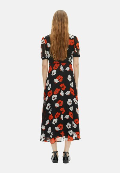 Frau mit langem Haar, die ein schwarzes Midikleid mit rot-weißen Blumenmuster trägt, steht mit dem Rücken zur Kamera und trägt schwarze Schuhe vor einem weißen Hintergrund.