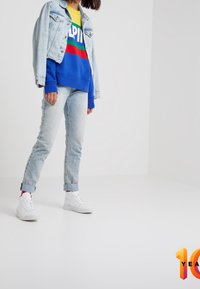 Blå sweatshirt med "ALPIN"-text, gul under, jeansjacka, ljusblå jeans och vita sneakers med röda detaljer. Enkel, avslappnad passform.