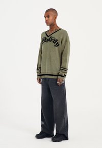Maglione a maglia khaki con dettagli in nero, caratterizzato da un scollo a V e maniche a righe. Abbinato a jeans neri larghi e sneakers.