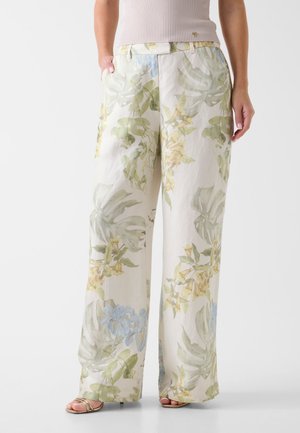 Pantaloni a gamba larga color crema con stampa floreale verde e gialla, indossati con top beige senza maniche a coste e sandali dorati con cinturini.