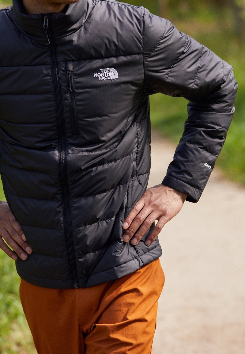 Black puffer jacket with a high collar, zip closure, and stitched panels. Features a logo on the chest and elastic cuffs. Paired with orange pants.