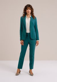 Komplet srajca in hlače iz teksturirane tkanine v modrozelena barva. Pod njo bela srajca. Oprijeta kroj z enim zapenjanjem na gumb. Črpalke s stiletto peto.