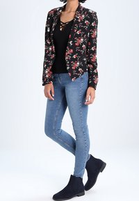 Veste noire à motif floral avec des fleurs rouges et blanches, portée sur un haut noir, associée à un jean skinny bleu clair et des bottines noires.