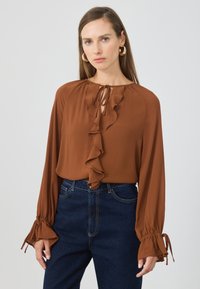 Kvinne iført en rustfarget bluse med frontruff og knyting, kombinert med mørkeblå høytlivsjeans og gullhockeyring.