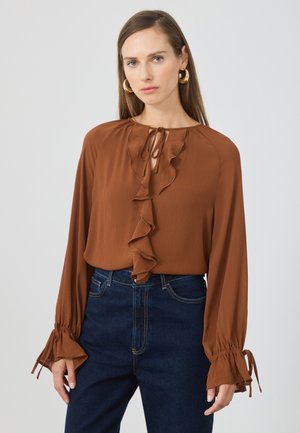 Woman wearing a rust-colored blouse with front ruffle and tie detail, paired with dark blue high-waisted jeans and gold hoop earrings.