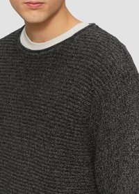 Donkergrijze gebreide sweater met een gestructureerd patroon, ronde halslijn, en een gelaagde witte blouse eronder. Close-up van de stof.