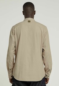 Beige långärmad skjorta med bakstycke, knappade ärmslut och rundad kant. Tillverkad av lättviktig tyg med en mjuk textur.