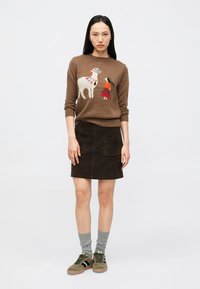 Pull en maille marron avec une finition texturée, présentant un motif coloré d'un chien et d'une fille. Elle porte une jupe en daim foncé et des baskets vertes.