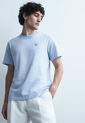 Jeune homme aux cheveux bouclés portant un t-shirt bleu clair avec un petit emblème en forme de cœur et un pantalon blanc, se tenant devant un fond clair uni.