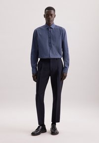 Uomo in piedi che indossa una camicia azzurra a fantasia con bottoni, pantaloni scuri su misura, calzini neri e scarpe eleganti nere su sfondo neutro.
