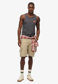 Camiseta sin mangas gris con pequeño logo rojo, pantalones cortos de cargo beige con bolsillos, camisa de franela a cuadros rojos y blancos atada a la cintura, zapatillas blancas de caña alta.