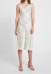 Robe midi fleurie en crème, fabriquée en tissu soyeux. Dotée d'un col en V et de détails froncés. Accentuée de petites fleurs vertes et bleues.