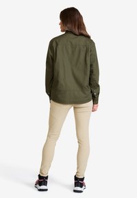 Kvinna som bär en olivgrön jacka, beiga åtsittande byxor och svarta och rosa sneakers, står vänd bort från kameran mot en vit bakgrund.