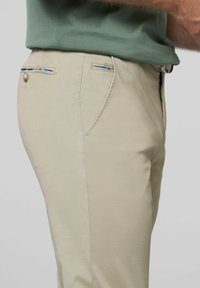 Beige broek van een textuurstof. Heeft accentstiksels en een achterzak met een decoratieve streepdetail.
