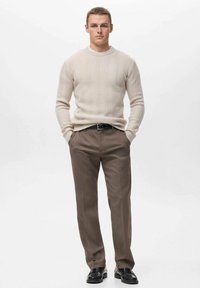 Suéter de punto trenzado beige claro, pantalones marrones ajustados y zapatos negros. El suéter presenta un cuello redondo y un patrón texturizado.