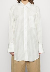 Chemise blanche à boutons réalisée en tissu léger, avec un motif en zigzag subtil, un col pointu et une poche poitrine unique. Manches longues.