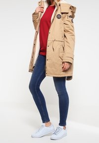 Beige parka-jas met een met bont gevoerde capuchon, voorzien van zijzakken, een trekkoord in de taille en geborduurde patches. Gedragen over een rode sweatshirt en blauwe jeans.